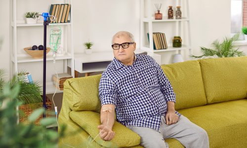 Elderly male patient getting intravenous treatment at home. Senior man sitting on couch with iv drip attached on his hand. Treatment, elderly healthcare, maintenance of immune system